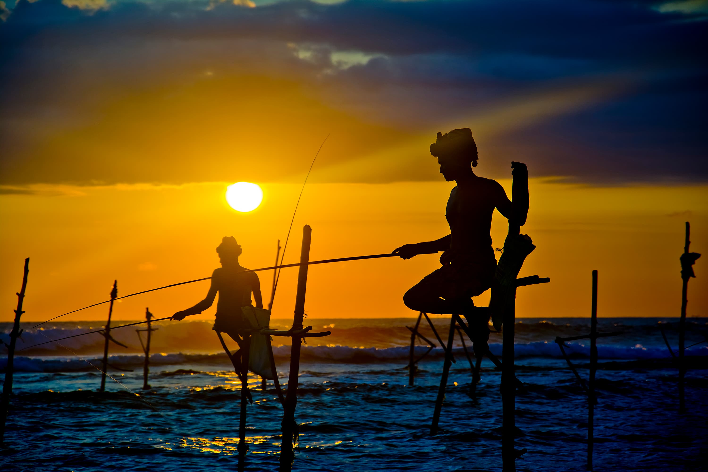 Silhouettes of traditional Sri Lankan stilt fishermen against a blazing golden sunset over the Indian Ocean