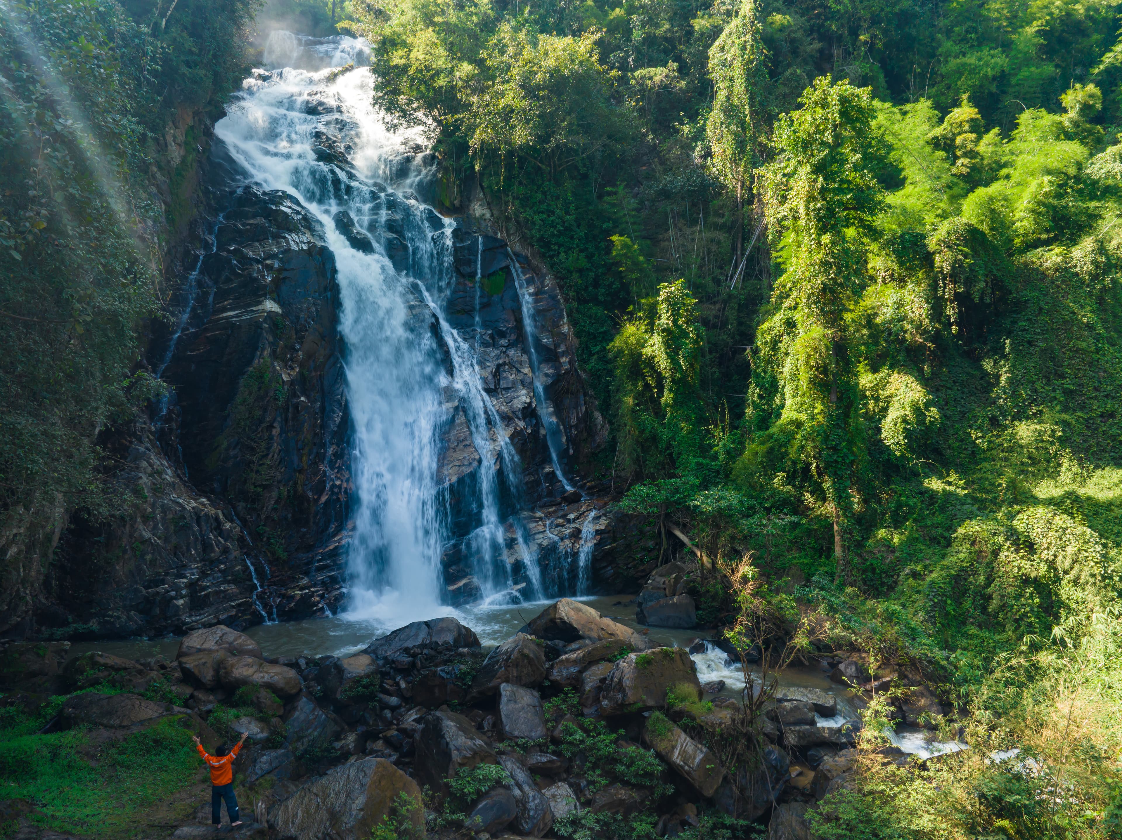 Adventurer standing with arms raised in triumph before a massive cascading waterfall in lush Sri Lanka jungle